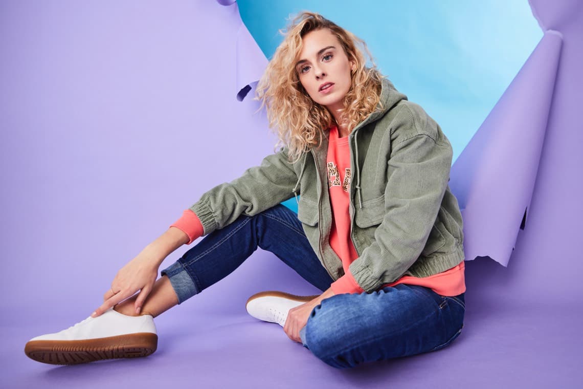 Frau mit lockigem Haar, trägt grüne Jacke, pinken Kapuzenpulli, Jeans und weiße Sneaker, sitzt vor lila und blauem Hintergrund