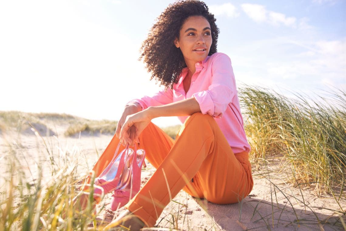 Person mit lockigem Haar sitzt im Sand, trägt pinkes Hemd und orange Hose, hält pinke Schuhe in der Hand, umgeben von Dünengras, sonniger Himmel.