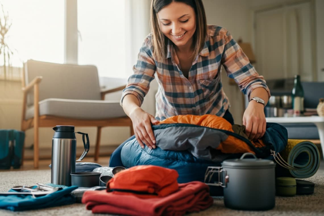 Eine Frau packt einen Schlafsack in einen Rucksack. Um sie herum liegen Campingutensilien, Kleidung und eine Thermoskanne auf dem Boden.