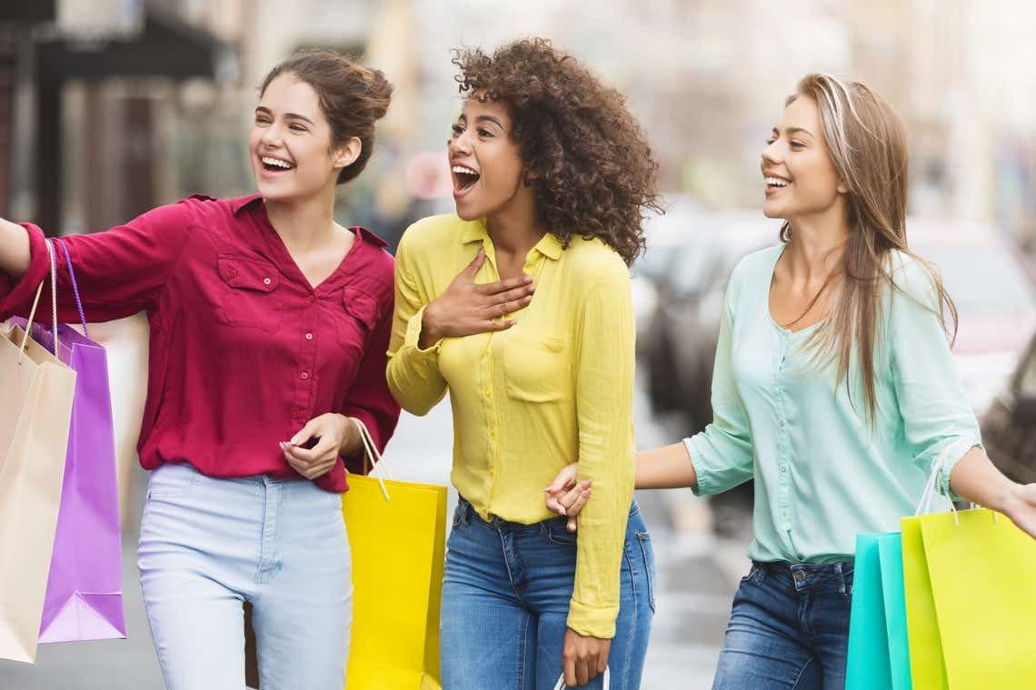 Drei Frauen gehen auf einer Straße in der Stadt, tragen rote, gelbe und mintgrüne Blusen.