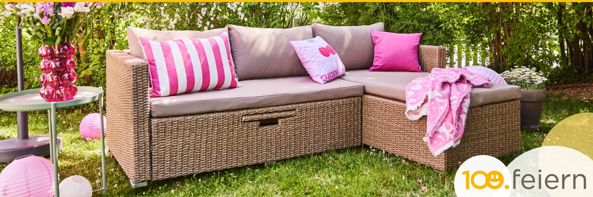 Loungesofa aus beigem Rattangeflecht, steht im Garten. Deko rosa-gestreifte Kissen.