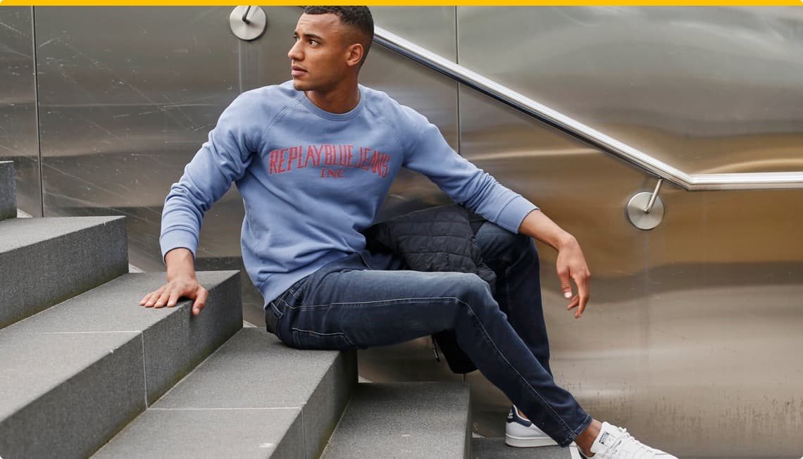 Mann sitzt auf Treppenstufen, trägt ein blaues Sweatshirt mit "REPLAY BLUE JEANS" und kombiniert es mit dunklen Jeans.