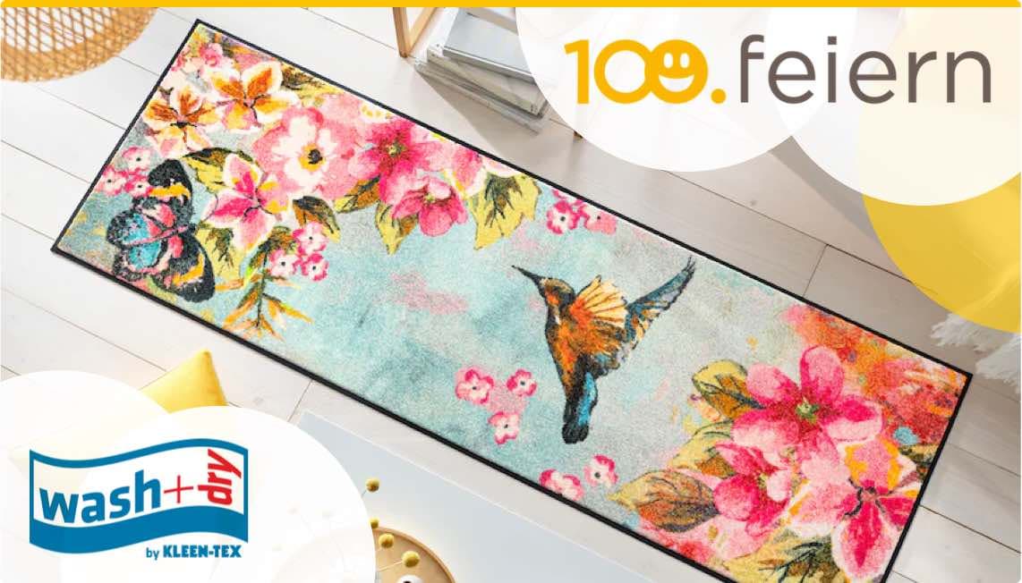 Bunter Teppich mit Blumen und einem Kolibri, beworben mit "100. feiern" und "wash+dry by KLEEN-TEX".