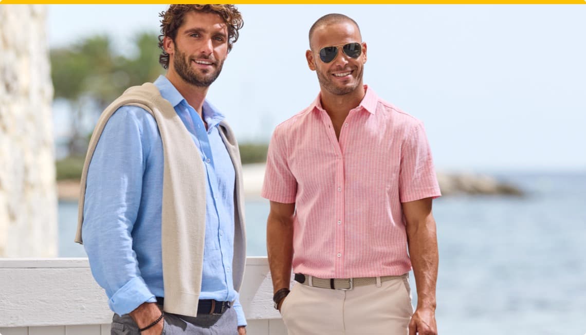 Zwei Männer stehen am Strand: einer in hellblauem Hemd mit Pullover, der andere in rosa kariertem Hemd, beide lächeln.