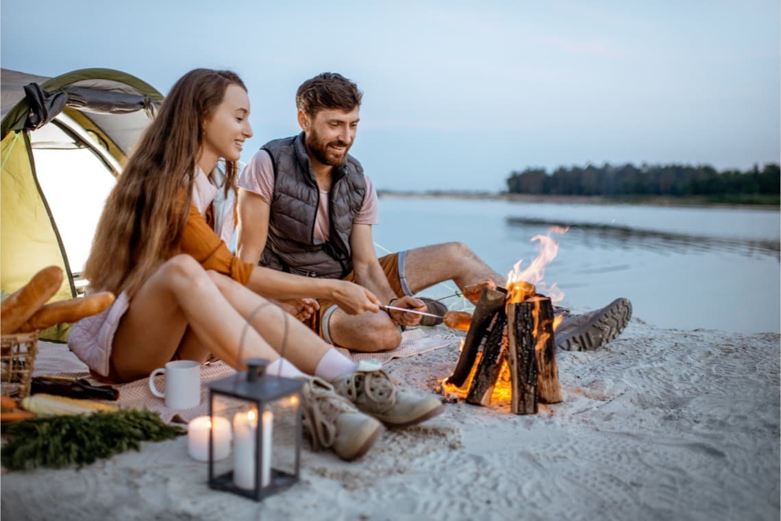 Paar sitzt am Strand vor ihrem Zelt am Lagerfeuer, romantische Abendstimmung.