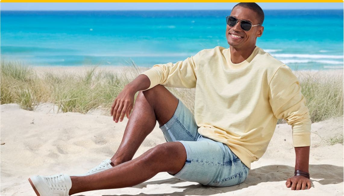  Mann in hellgelbem Sweatshirt und Jeansshorts sitzt entspannt am Strand.