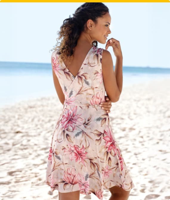 Leichtes, blumiges Kleid mit V-Ausschnitt am Rücken, getragen am Strand, umgeben von feinem Sand und Meer. 