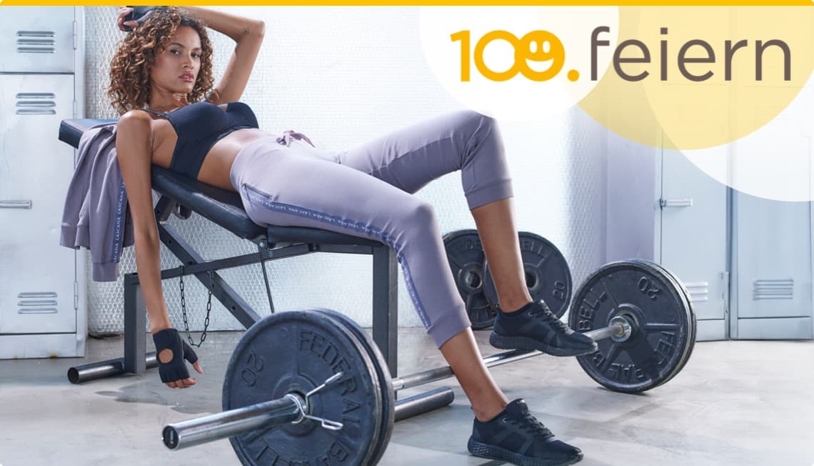 100. feiern – Frau in Sportkleidung liegt auf einer Hantelbank im Fitnessstudio, Hanteln auf dem Boden