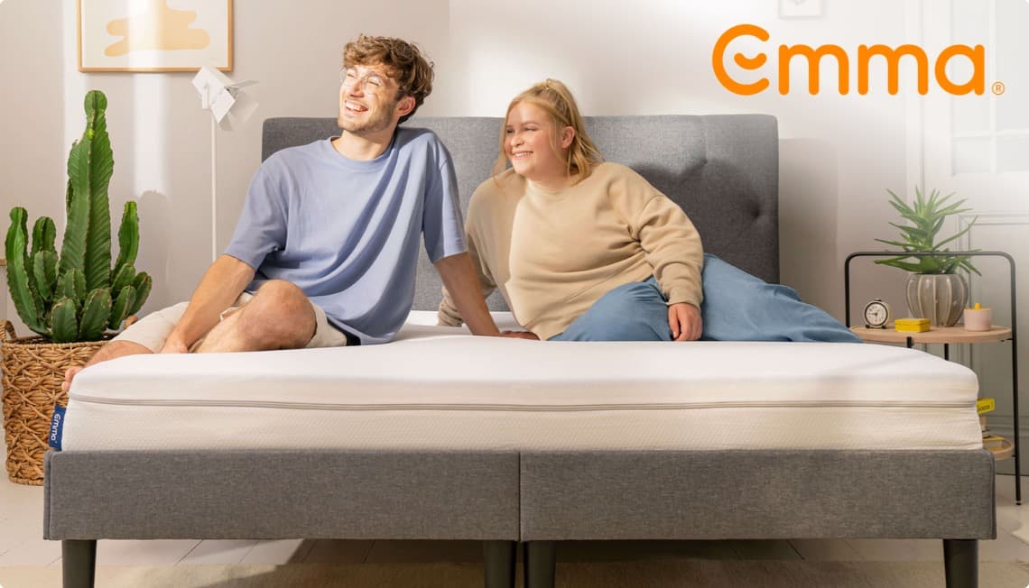 Emma – Mann und Frau sitzen entspannt auf einem Bett mit einer Emma-Matratze, Zimmerpflanze und Nachttisch im Hintergrund