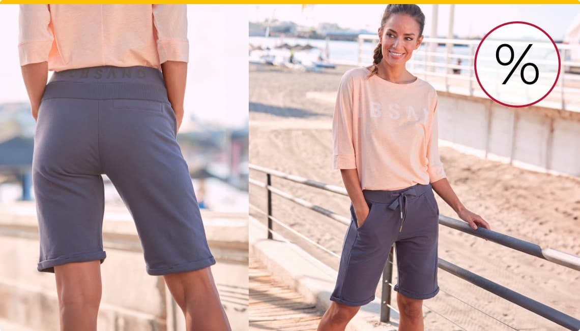 Prozentzeichen – Frau am Strand trägt helles Shirt und dunkle Shorts, Hände in den Taschen, Blick aufs Meer