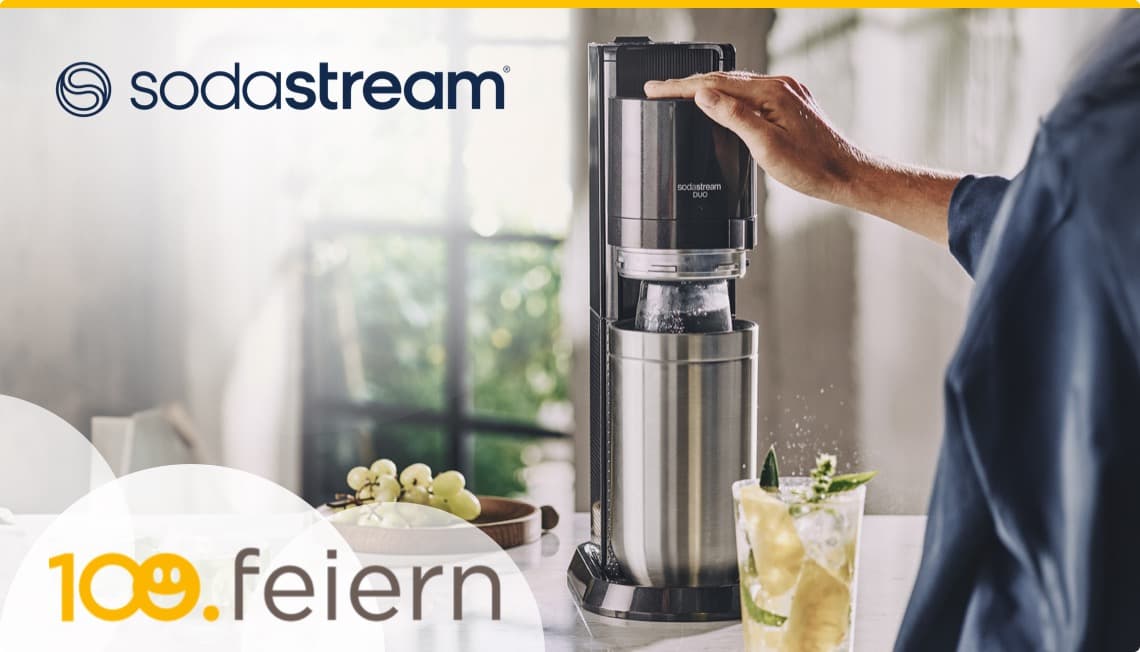 Sodastream 100. feiern – Person bedient Sodastream DUO neben Glas mit Getränk und Trauben auf Küchentisch