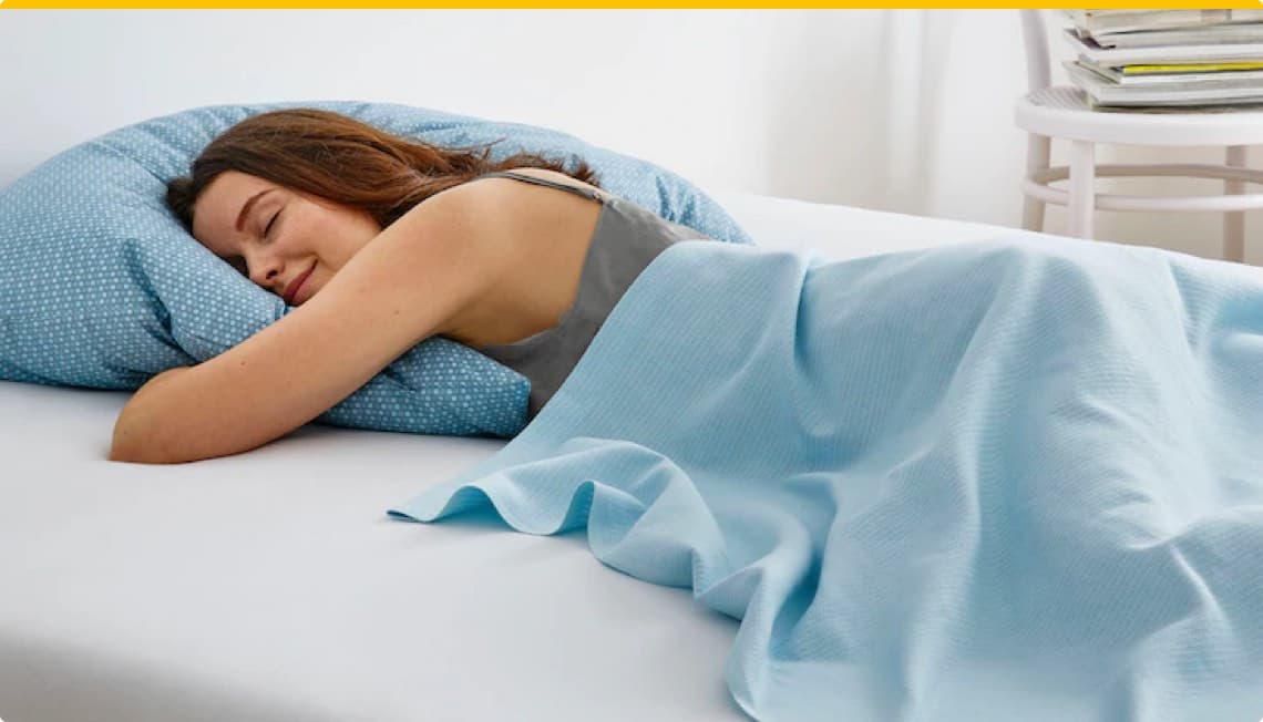 Frau liegt auf dem Bauch und schläft, zugedeckt mit einer hellblauen Decke auf einem Bett mit blau gepunktetem Kissen.
