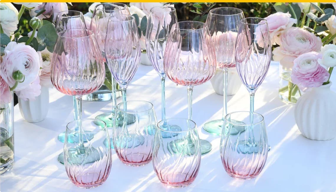 Mehrere rosa und blaue Weingläser auf einem Tisch, umgeben von weißen und rosa Blumen in Vasen