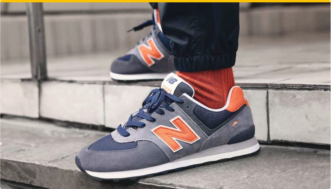 Graue New Balance Sneaker mit orangefarbenem Logo und roten Socken auf einer Treppe.