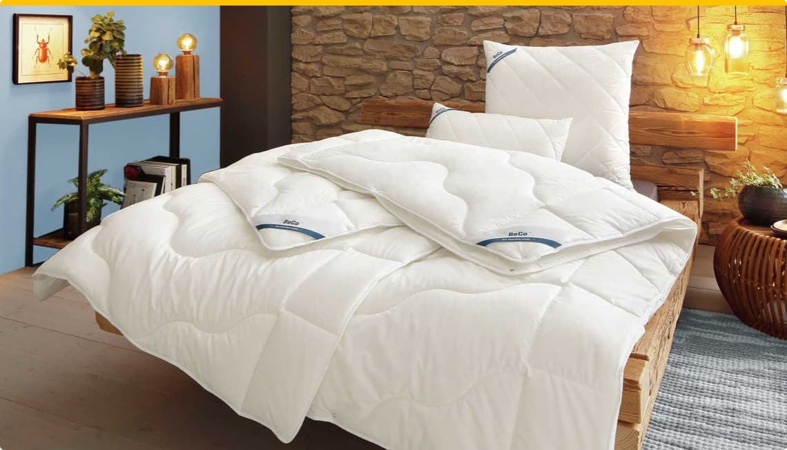 BeCo – weißes Bettenset bestehend aus Bettdecke und Kissen auf Holzrahmenbett vor Steinwand und blauer Wand mit Regal