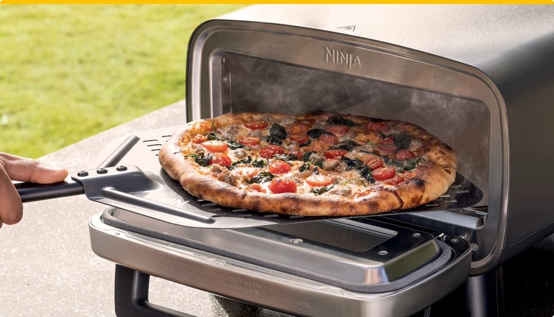 Ninja Pizzaofen – Hand zieht frische, dampfende Pizza mit Tomaten und Spinat aus dem Ofen auf einer Schaufel