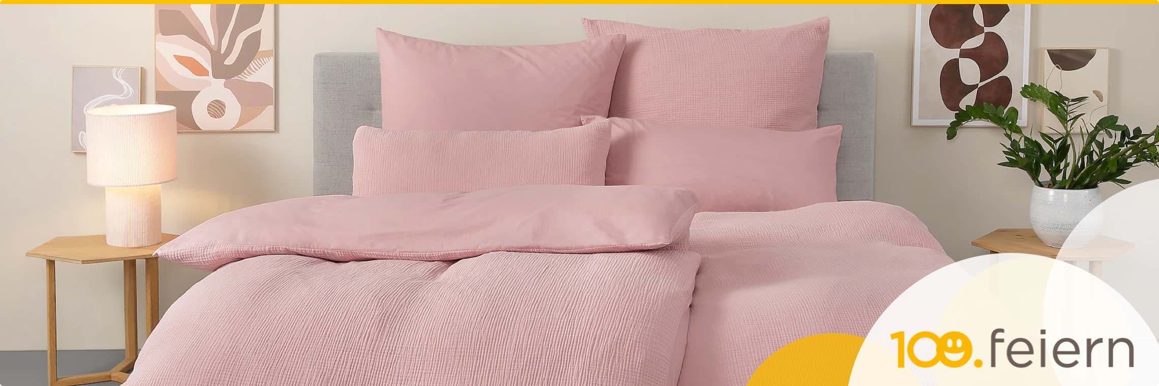 100. feiern – Schlafzimmer mit rosa Bettwäsche, grauem Kopfteil, Nachttisch, Lampe, Pflanzen und abstrakten Bildern an der Wand
