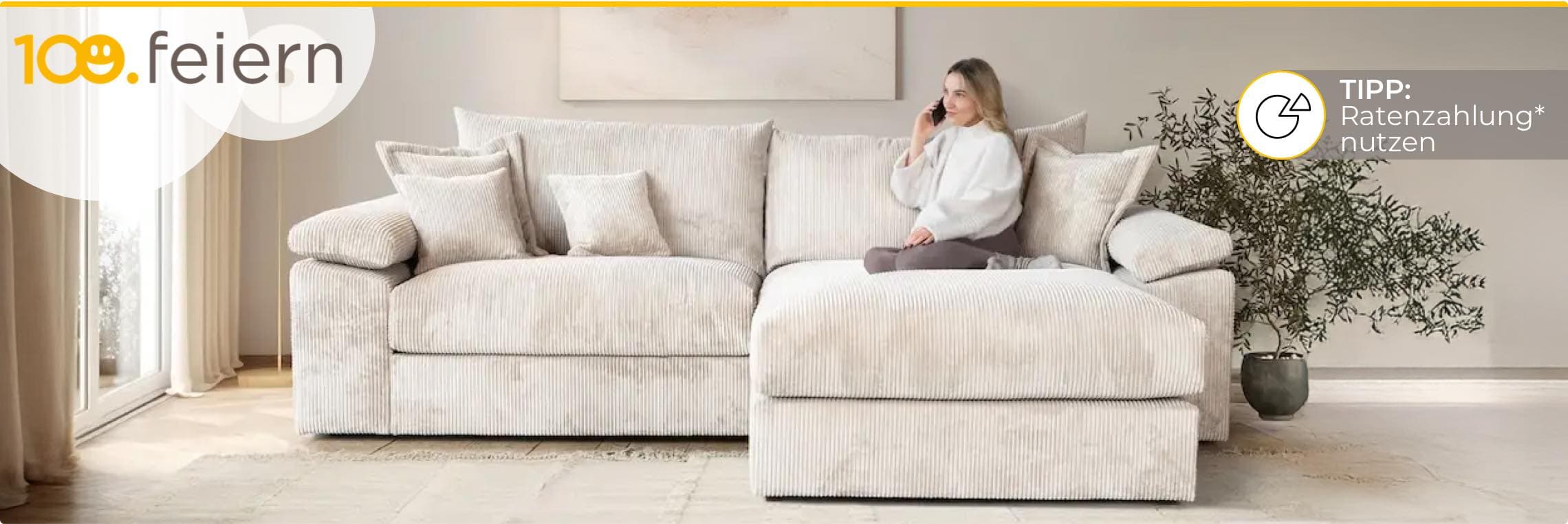 100 feiern – Tipp: Ratenzahlung* nutzen – Frau sitzt auf großer, beiger Couch in modernem Wohnzimmer