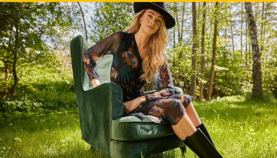 Frau in transparentem Blumenkleid und Hut sitzt auf grünem Sessel im Wald