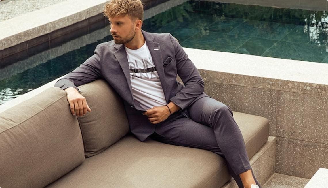 Mann in grauem Anzug und weißem T-Shirt sitzt entspannt auf einem Sofa neben einem Pool