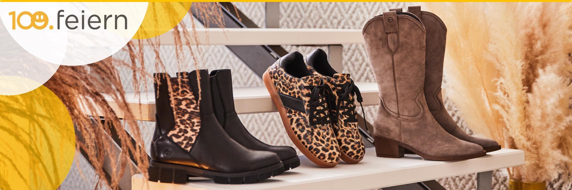 100. feiern – Drei Paar Schuhe auf einem Regal: schwarze Stiefeletten mit Leopardenmuster, Sneaker mit Leopardenmuster, braune Stiefel