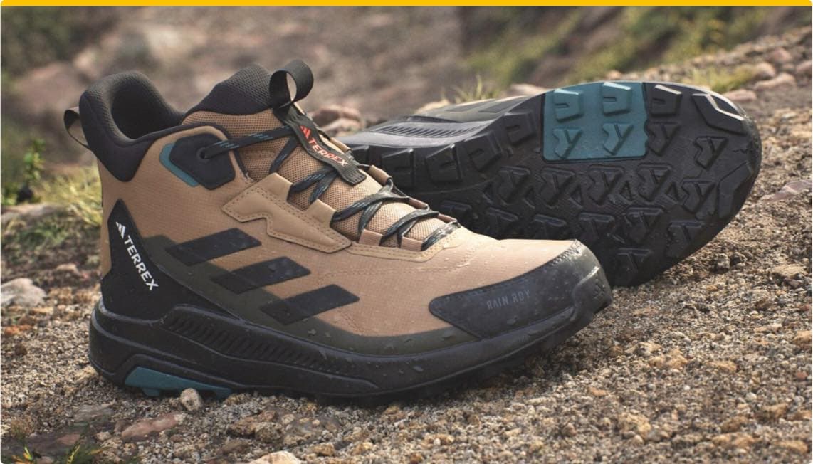 Adidas Terrex Wanderschuhe in Braun und Schwarz auf sandigem Boden, einer steht aufrecht, der andere liegt mit der Sohle nach oben.