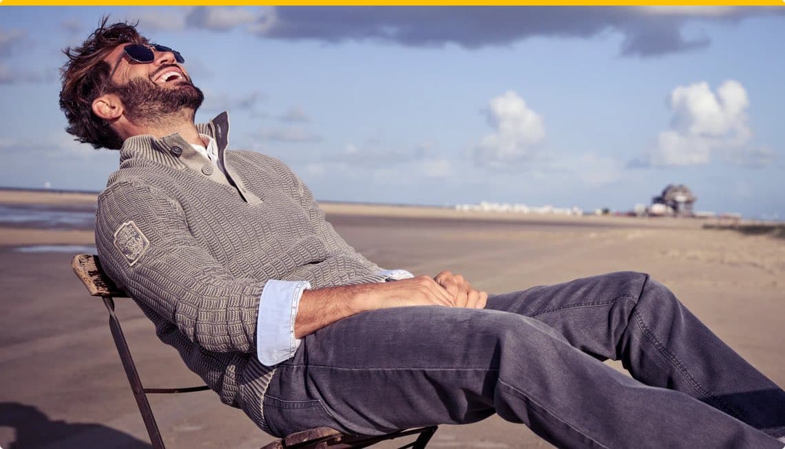 Mann in grauem Pullover und Jeans lehnt entspannt in einem Stuhl am Strand