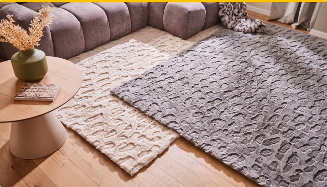 Beige und graue Teppiche mit Muster, braunes Sofa und runder Holztisch mit grüner Vase und Buch darauf in gemütlichem Wohnzimmer