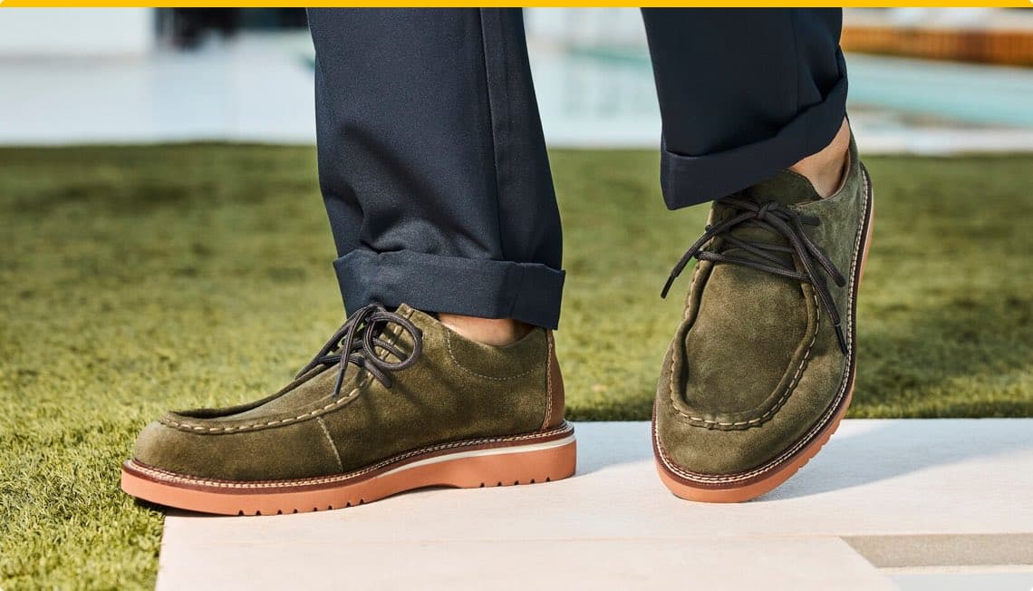 Person trägt olivgrüne Schnürschuhe und dunkelblaue Hosen, steht auf einem grünen Rasenteppich.