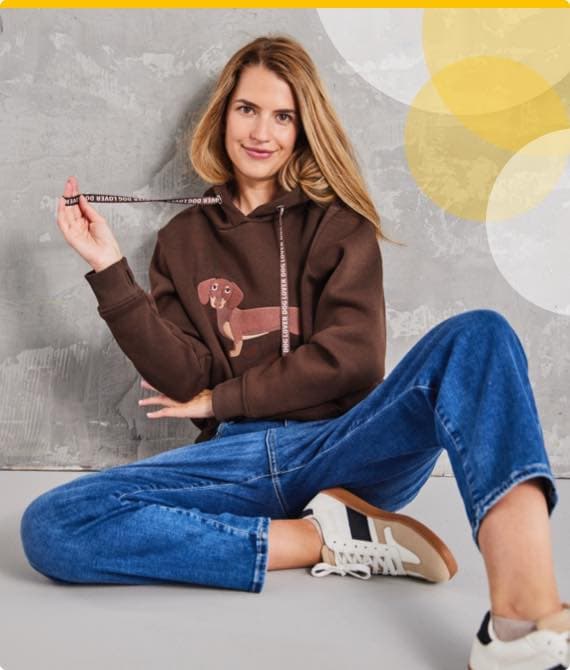 Frau in braunem Kapuzenpullover mit Hundemotiv und blauer Jeans sitzt am Boden und hält die Kordel des Hoodies