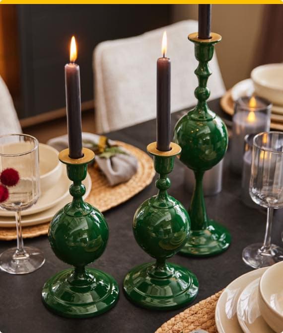 Drei grüne Kerzenständer mit brennenden schwarzen Kerzen auf einem gedeckten Tisch mit Gläsern und Tellern.