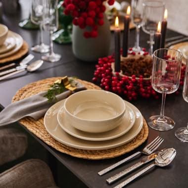 Festlich gedeckter Tisch mit Tellern, Besteck, Gläsern und Weihnachtsdeko mit roten Beeren und Kerzen im Hintergrund