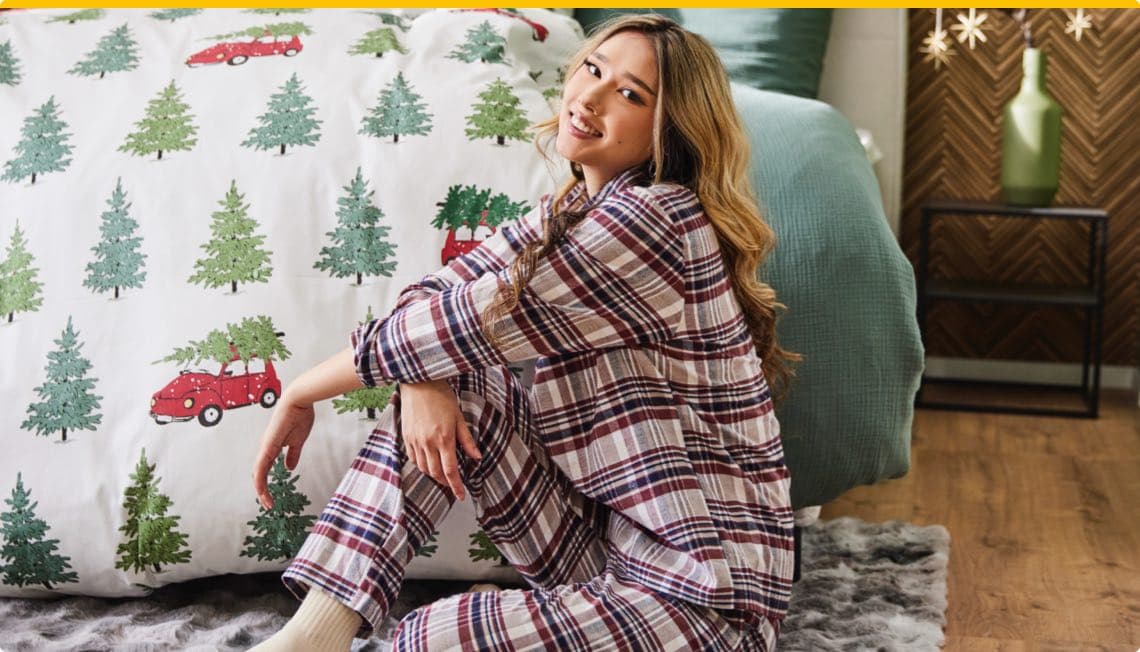 Frau in kariertem Pyjama sitzt vor Bett mit Weihnachtsbettwäsche