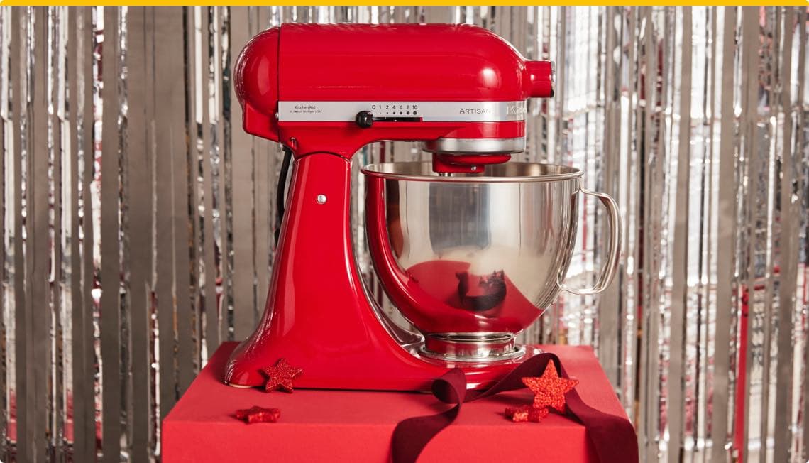 Rot glänzende Küchenmaschine mit silberner Rührschüssel, dekoriert mit roten Glitzersternen vor silbernem Vorhang.