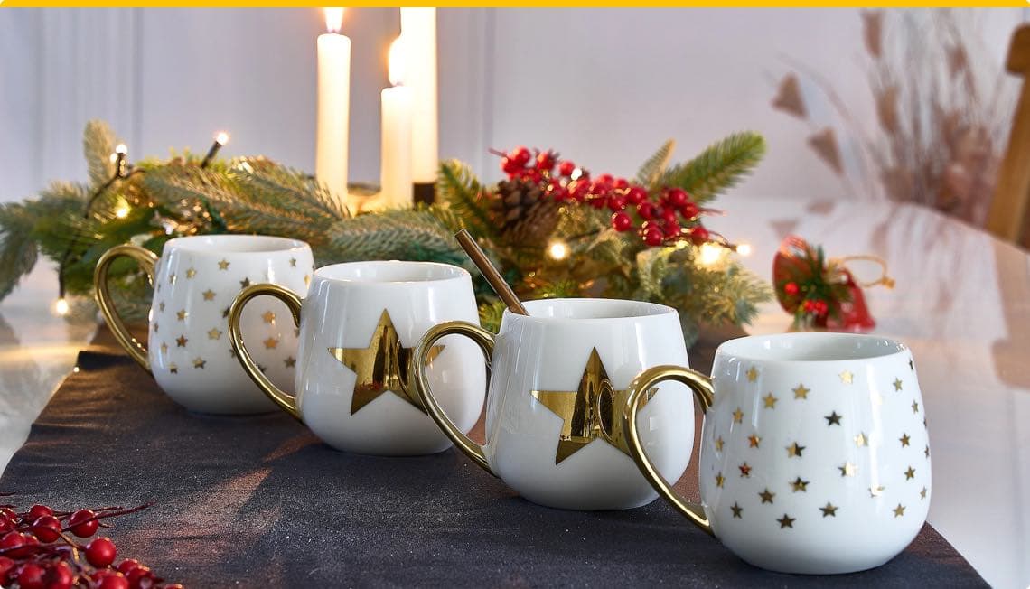 Vier weiße Tassen mit goldenen Sternen vor Weihnachtsdekoration mit Kerzen und Tannenzweigen