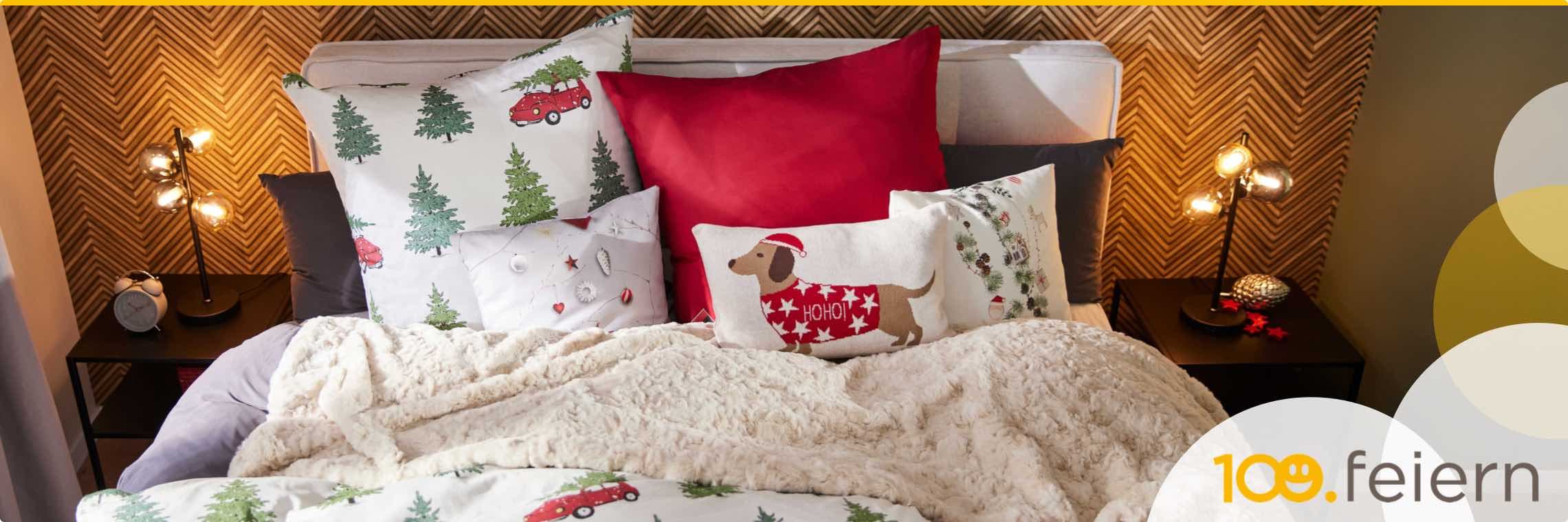 109.feiern – gemütliches Bett mit Weihnachtskissen, Hund in rotem Pullover, Tannenbaum- und Auto-Motiv, Nachttisch mit Lampe und Uhr