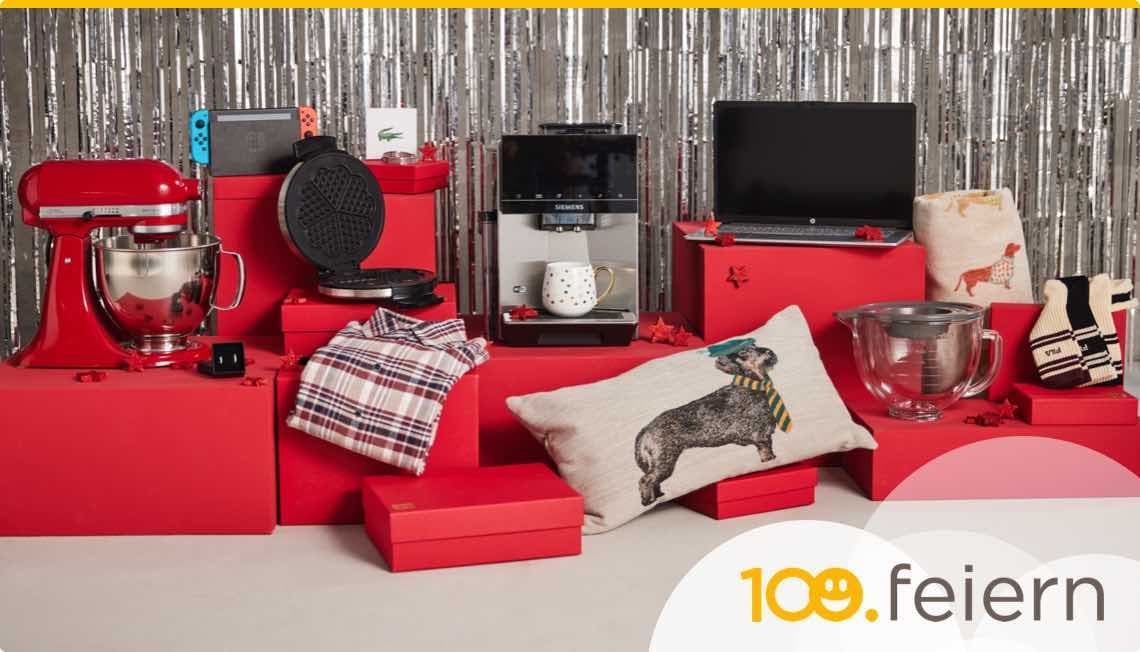 100. feiern – Rote Küchenmaschine, Waffeleisen, Laptop, Kissen und Kleidung auf roten Boxen vor silbernem Hintergrund