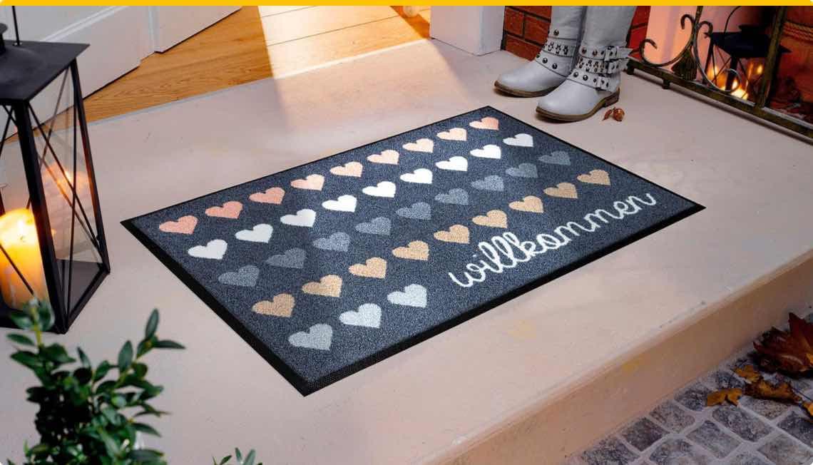 Willkommen – Fußmatte mit Herzmustern auf einer Veranda, daneben stehen weiße Stiefel und eine Laterne mit Kerze.