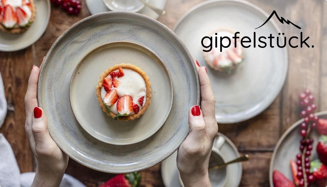 gipfelstück – Hände halten Teller mit Erdbeertörtchen, weitere Törtchen im Hintergrund auf Holztisch