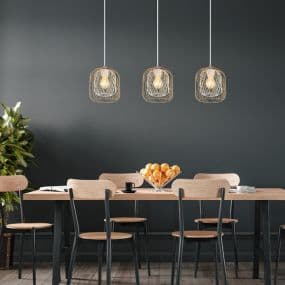 Drei hängende Lampen über einem Esstisch mit Holzstühlen und einer Schale mit Orangen. Pflanze links im Hintergrund.