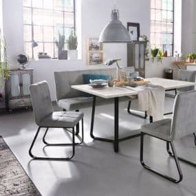 Moderner Essbereich mit Tisch, grauen Stühlen und Bank vor großen Fenstern. Einrichtungsgegenstände und Pflanzen im Hintergrund.