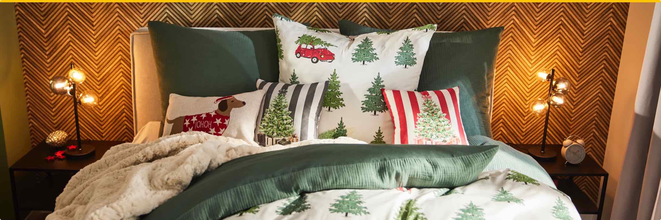 Bett mit Weihnachtskissen und Decken, dekoriert mit Tannenbaum- und Hundemotiven, daneben Nachttisch mit Lampe und Wecker.