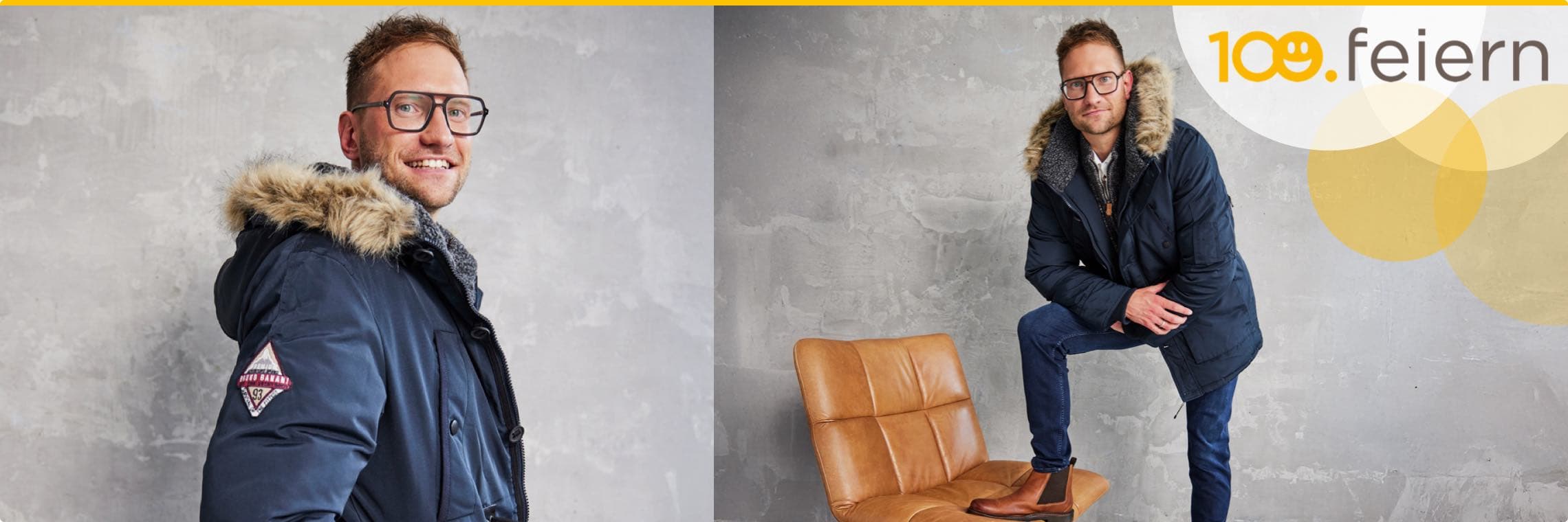 100. feiern – Mann in dunkler Winterjacke mit Fellkragen posiert vor grauer Wand und braunem Stuhl