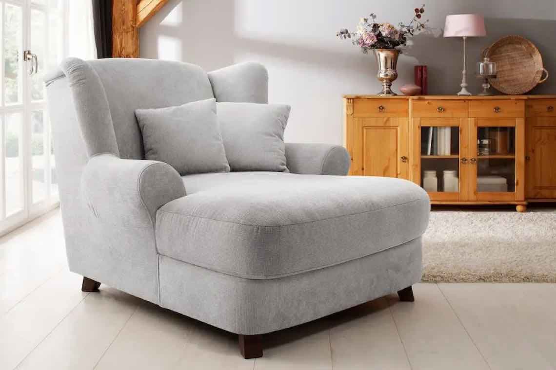 Grauer Sessel mit zwei Kissen, Holzkommode mit Dekoration im Hintergrund, großer Fensterbereich auf der linken Seite