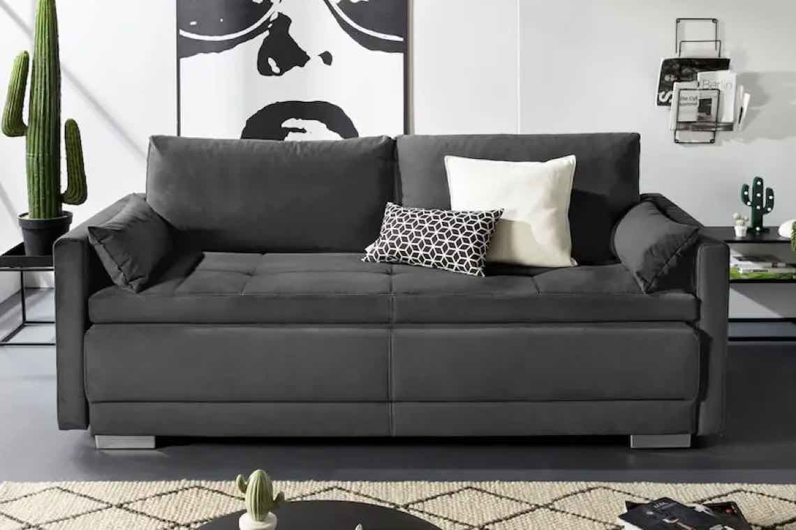 Dunkelgraues Sofa mit zwei Kissen, eins weiß und eins schwarz-weiß gemustert, vor einer Wand mit großem Gesichtsposter