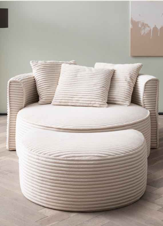 Rundes, beiges Sofa mit gerippten Kissen und passendem Hocker auf hellem Holzboden, Leinwand an der Wand im Hintergrund