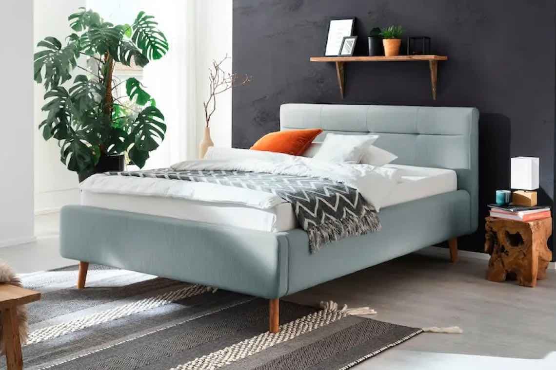 Modernes Schlafzimmer mit grauem Polsterbett, orangefarbenem Kissen, Pflanze, Wandregal und Holztisch mit Büchern und Lampe