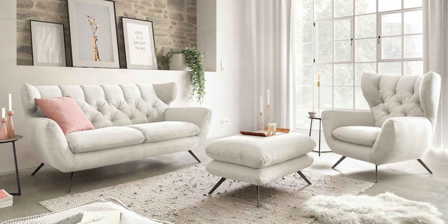 Modernes Wohnzimmer mit weißer Couch, Sessel, Hocker und rosa Kissen. Bilder an der Wand, große Fenster, Teppich und Kerzenständer.