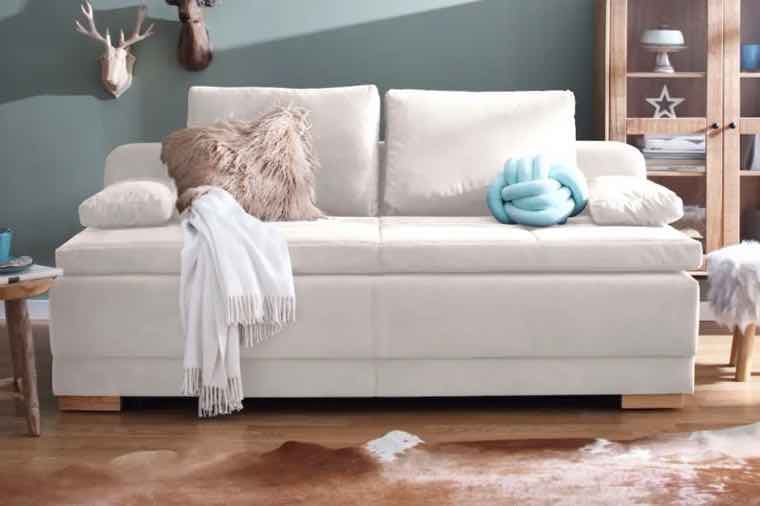 Weißes Sofa mit Kissen und Decken. Wanddeko mit Hirschkopf, Holzmöbel und Teppich im Hintergrund.