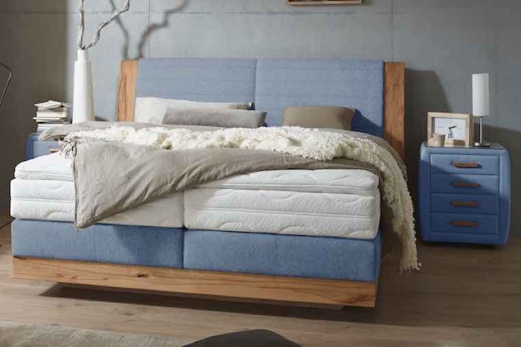 Doppelbett mit blauen Kopfteil, Decken und Kissen, daneben blauer Nachttisch mit Lampe, Bilderrahmen und Büchern